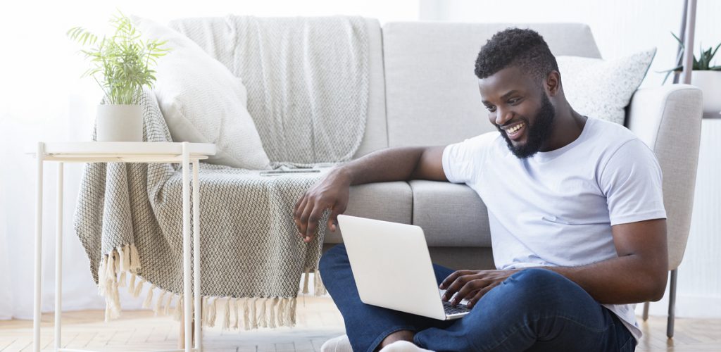 African guy playing video game on laptop computer, sitting on floor at home, free space