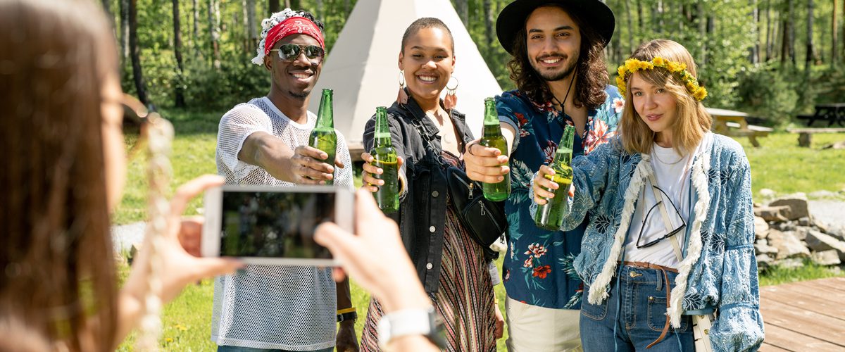 Group of positive young multi-ethnic people in hippie outfits posing with beer bottles for photo at countryside festival