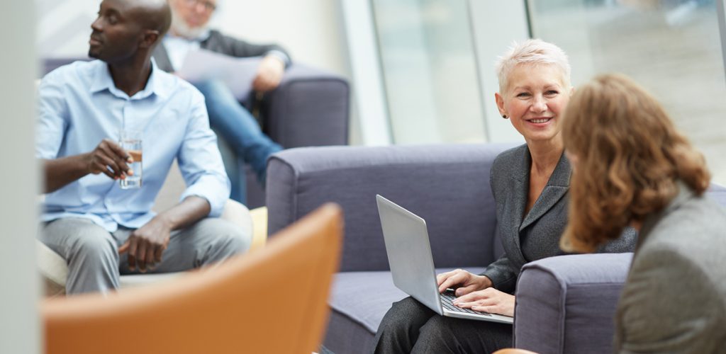 Portrait of successful senior businesswoman smiling cheerfully while talking to colleagues and using laptop during meeting in office hall, copy space
